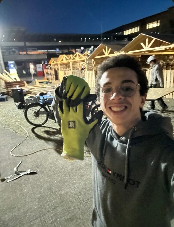 A student in glasses holds up a work glove in front of half-built wooden chalets at dusk.