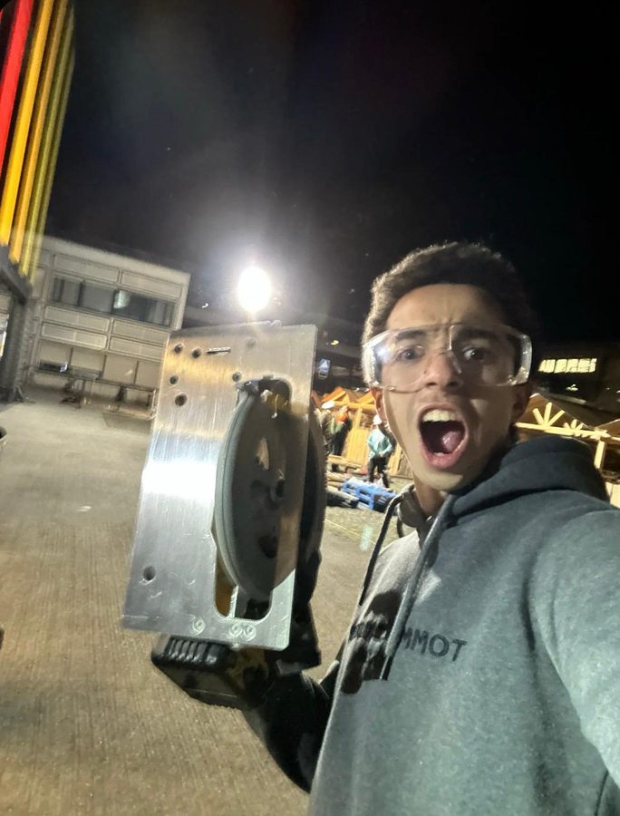 A student in safety glasses cheers at the camera, holding up a power tool on a building site at night.