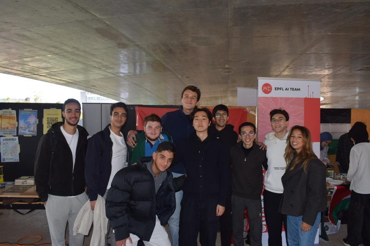 About fifteen students posing together in front of an EPFL AI Team stand under a concrete ceiling.