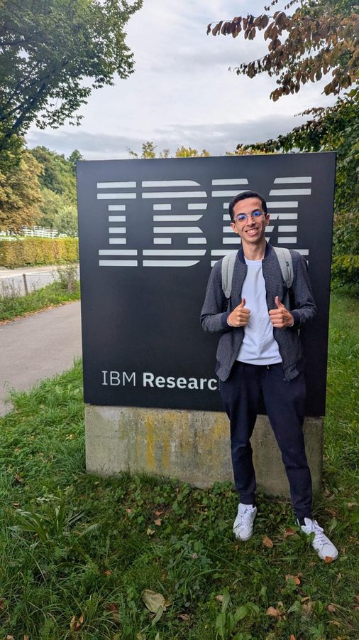 A student giving two thumbs up beside the IBM Research sign on a lawn.