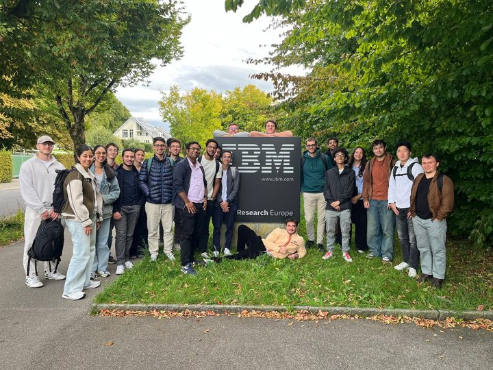 About twenty-five students posing around the IBM Research Europe sign among trees.