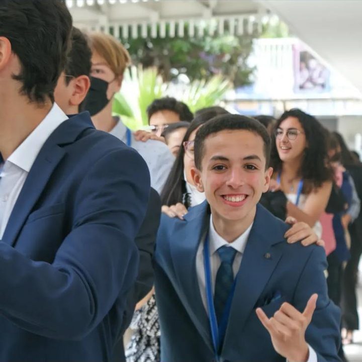 A grinning delegate in a navy suit making a shaka sign among other delegates.