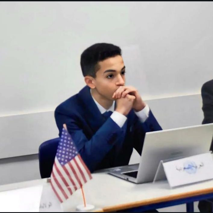 A delegate in a navy suit at a desk behind a small US flag and a laptop, hands clasped.