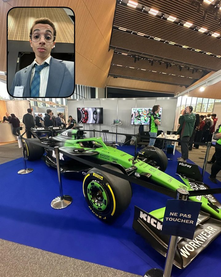 A green and black Formula 1 car on a blue carpet at a fair stand; an inset shows a student in a suit and glasses.