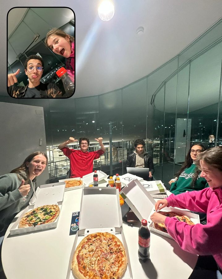 Students around a round table covered in pizza boxes in a glass-walled room at night, one cheering with both arms up; an inset shows two of them waving.