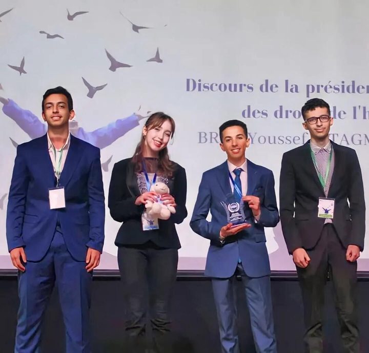 Four Model UN delegates on stage, Omar in a blue suit holding a glass award