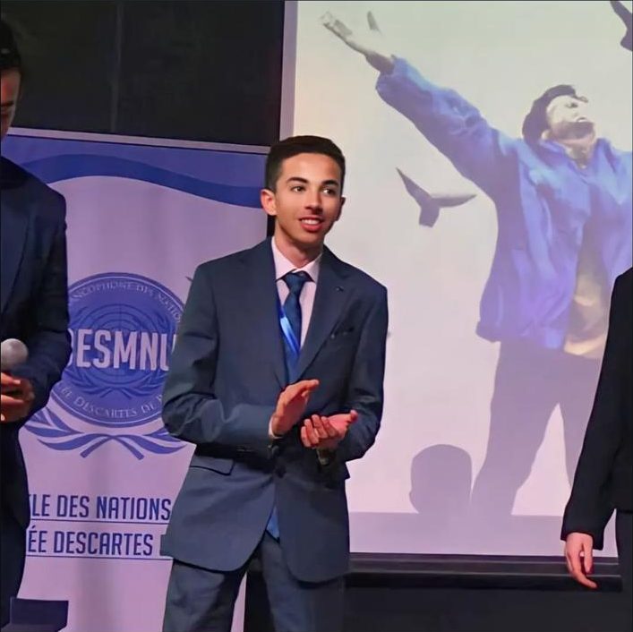 A delegate in a navy suit applauding on a stage, in front of a DESMNU banner and a screen.