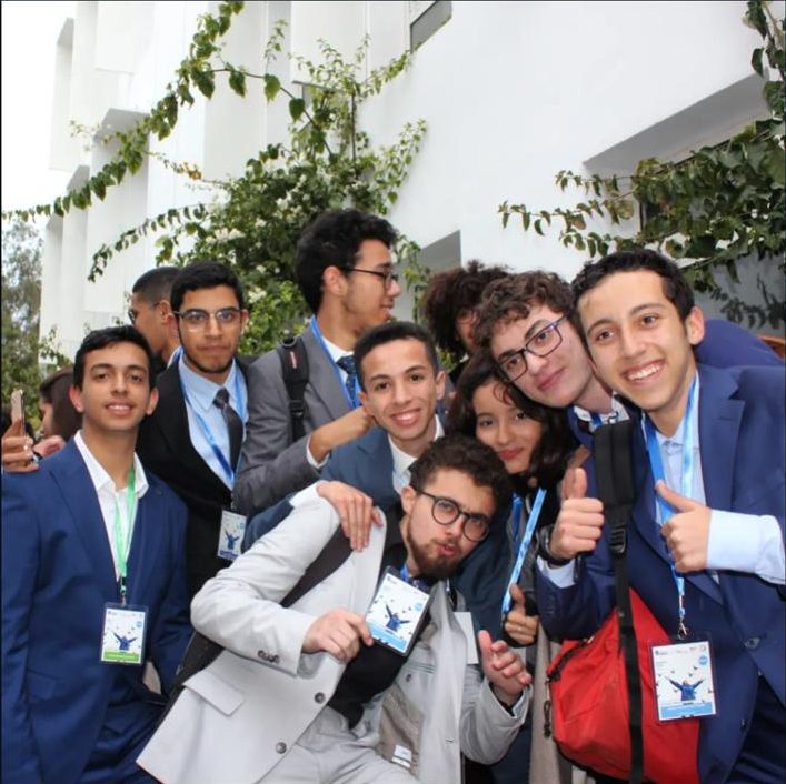 A crowd of delegates in suits squeezed into a selfie outdoors, several giving a thumbs up.