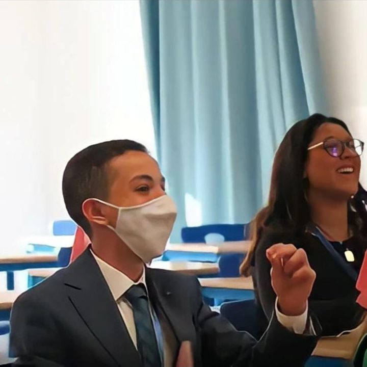 A masked delegate in a suit beside another who is laughing, in a committee room.