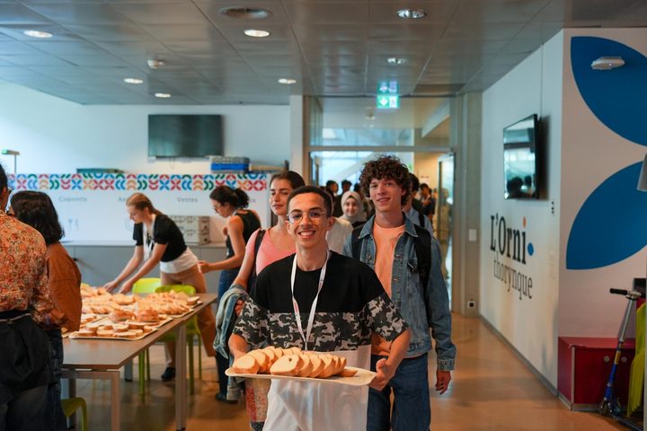 Students in aprons and lanyards carrying trays of bread through a canteen, one smiling at the camera.